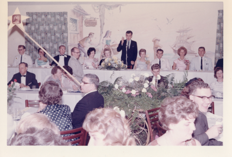 Man rises to give speech from head table while others are seated
