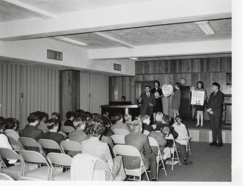 A man plays piano, women hold signs with song lyrics, and others sing and clap for a seated child audience
