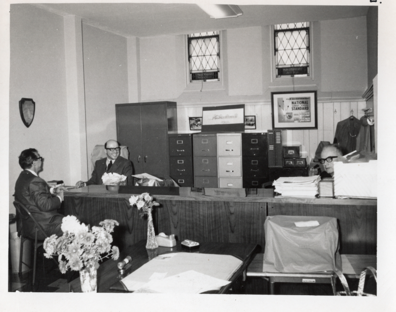 Three men sit at a table together in front of filing cabinets