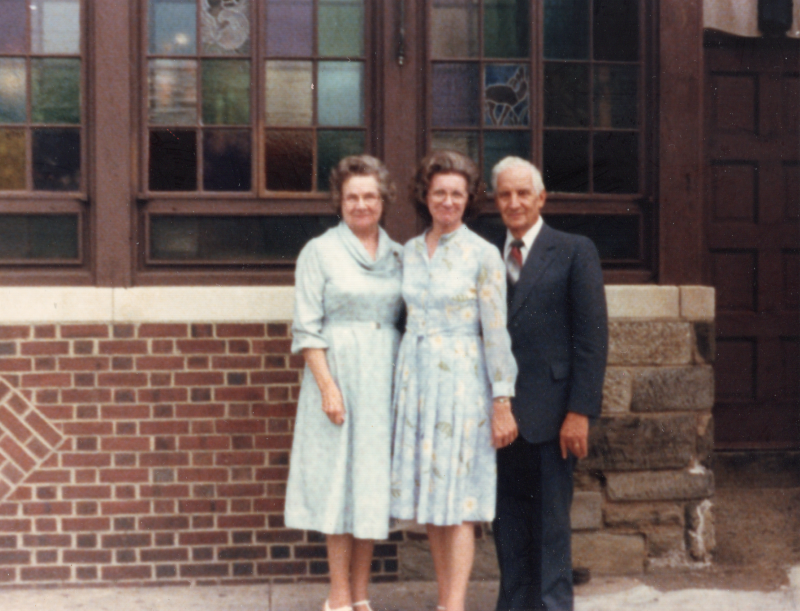 Two women and a man are photgraphed outside of a brick building with stained glass windows