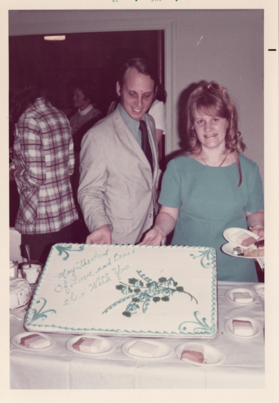 Man and woman pose with a cake that reads "May the Lord of Love and Peace Be With You" 