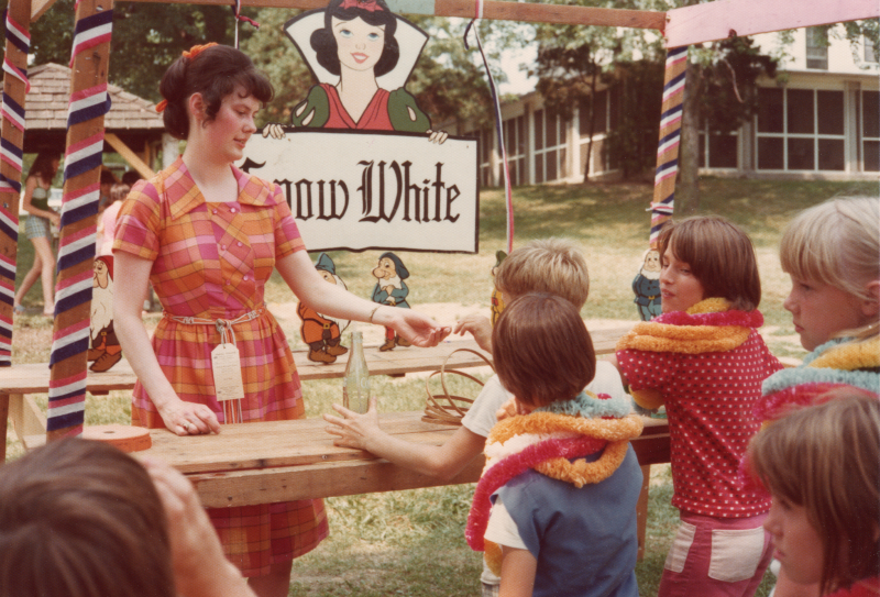 A woman speaks to a group of children who are wearing leis in an outdoor wood structure decorated with streamers and a portrait of Snow White