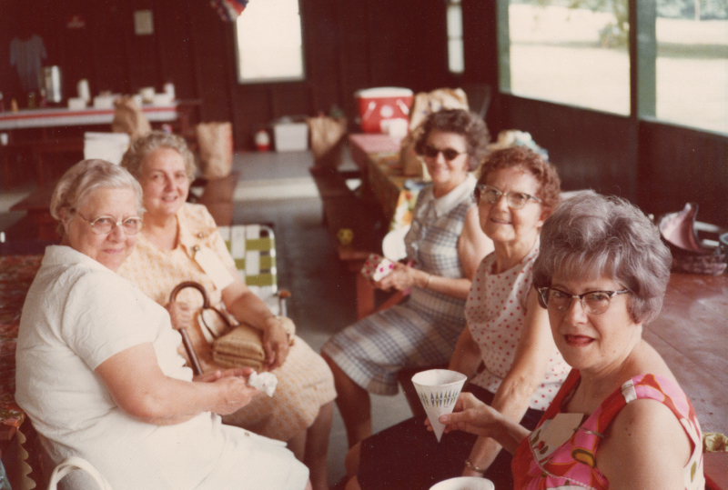 A group of older women are seated indoors enjoying refreshments