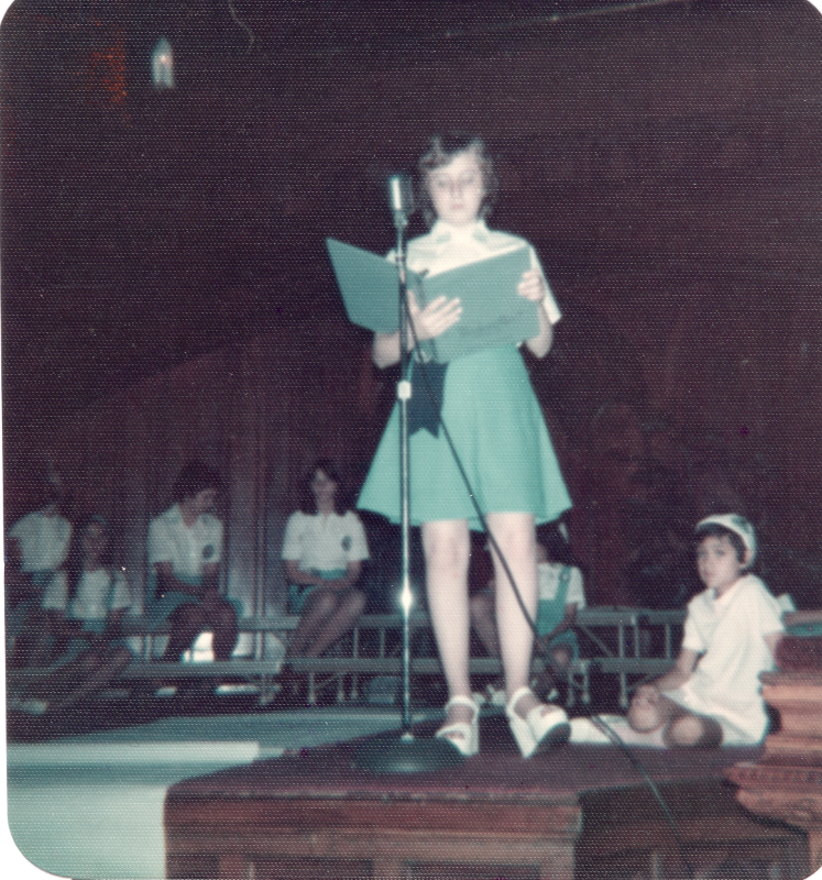 A missionette stands on stage in front of a microphone holding a book