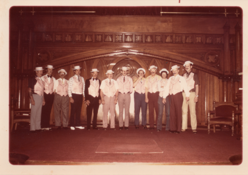 A line of men dress in the style of a barbershop quartet with red and white vertical stripes and fake mustaches