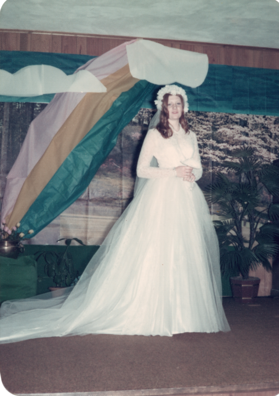 A woman in a wedding gown and veil speaks in front of a forest and rainbow backdrop