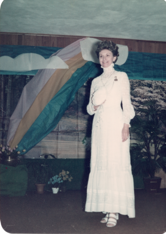 A woman in a wedding gown and veil speaks in front of a forest and rainbow backdrop