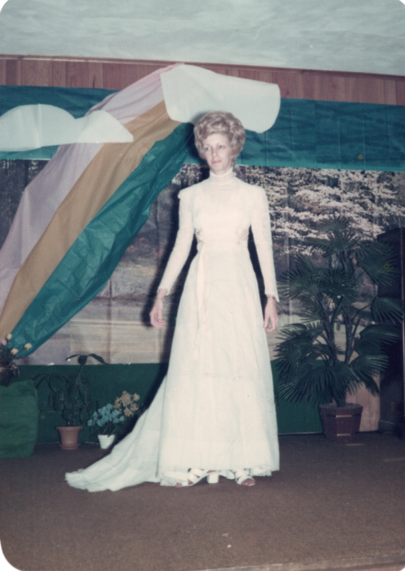 A woman in a wedding gown and veil stands in front of a forest and rainbow backdrop