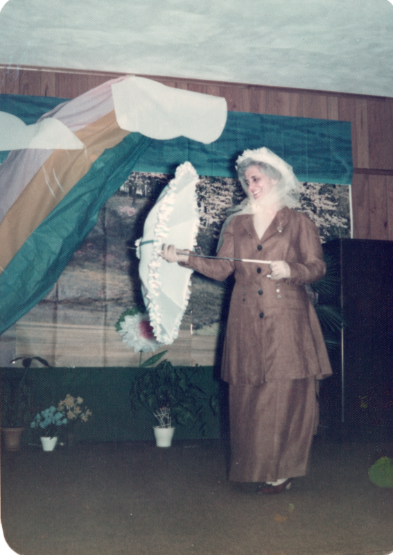 A woman in a tan suit with a white parasol stands in front of a forest and rainbow backdrop