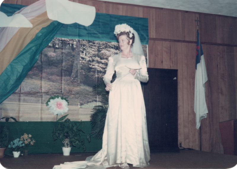 A woman in a wedding gown and veil reads from a paper in front of a forestal background and a rainbow