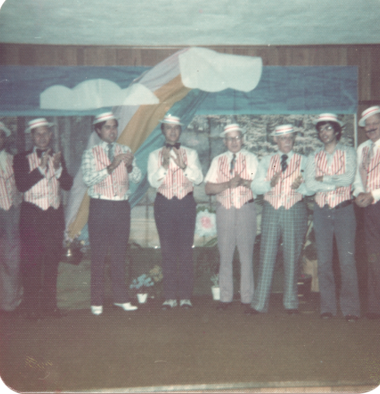 A group of men dressed in barbershop style perform in front of a forestal backdrop and a rainbow
