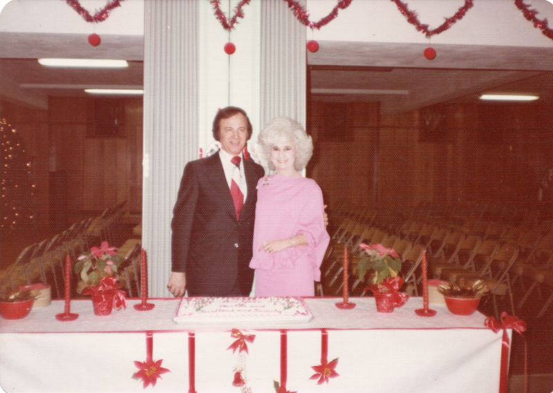 A man and woman stand with a cake in front of a Christmas tree