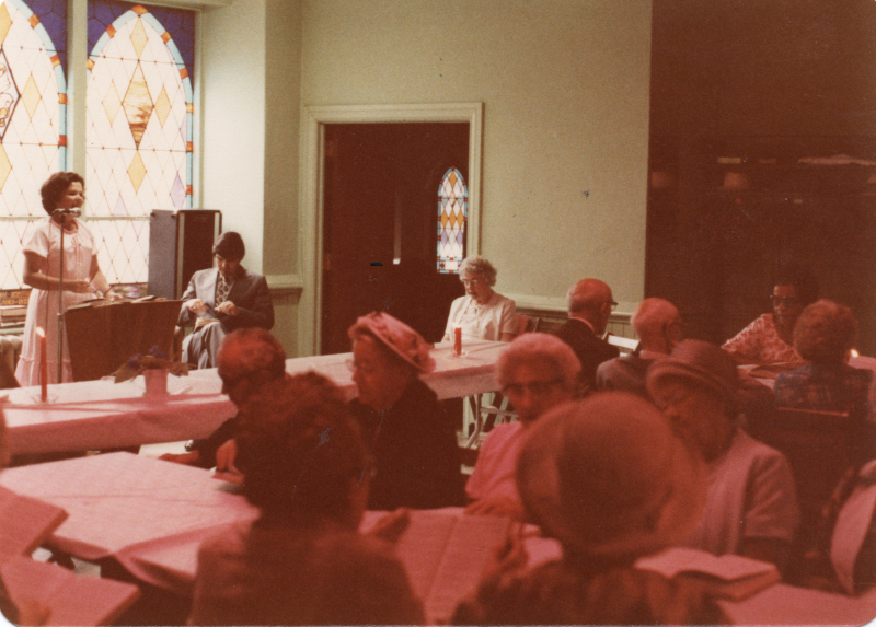 Mrs. Stone leads a song from a hymnal with people around tables with hymnals singing along