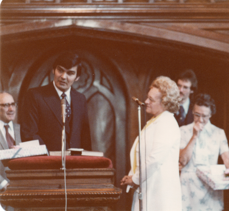 Pastor Stone speaks in the sanctuary, and is surrounded by congregants carrying a present