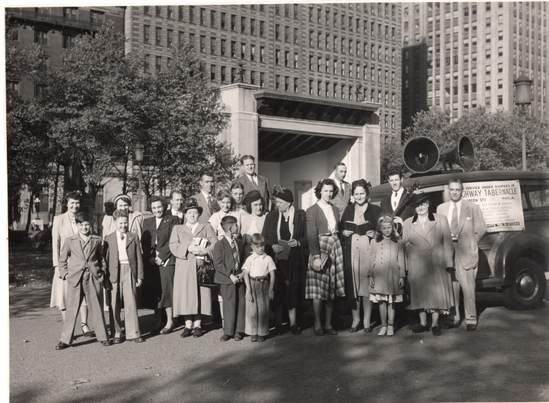 Men, women, and children carrying hymnals, Bibles, and instruments pose for a photograph outside with a car that invites the community to Highway Tabernacle