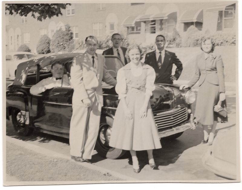 A group of three men and three women pose for a portrait with a car