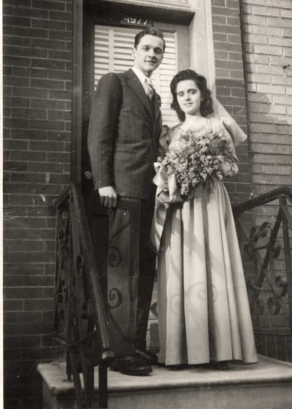A man stands with a woman in a wedding dress and veil in front of an outside door with the house number "1977"