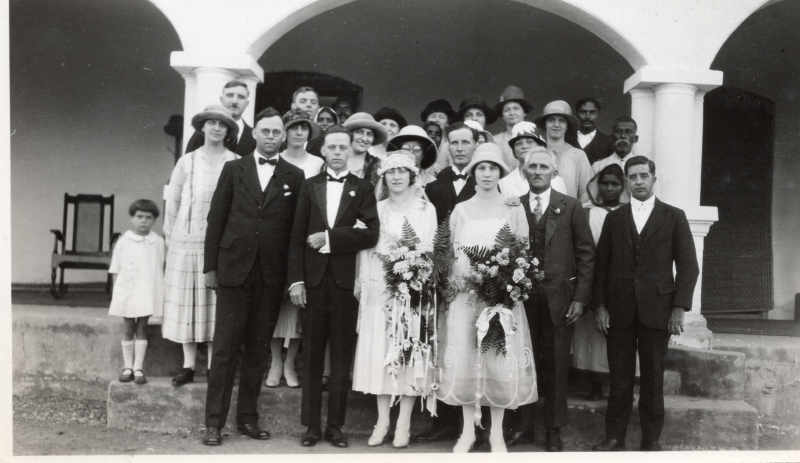 Portrait of a group dressed in formal clothing, potentially with two brides in white dresses and with flowers in the picture's center