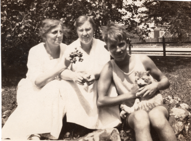Two women, a boy, and a cat smile together on a picnic blanket. Likely from the 08/1931 missionary rally at Marantha Park