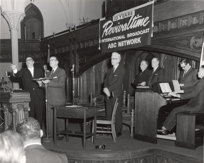 Radio broadcast of Revivaltime with two men singing into a microphone, three men standing in the background, and an organist in the sanctuary at Highway (18th and Spring Garden) in front of a Revivaltime banner