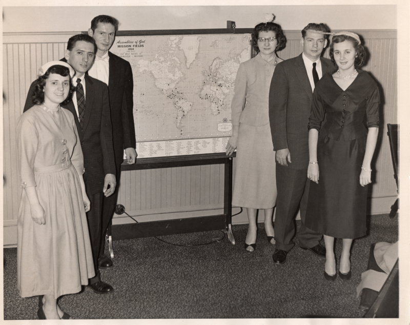 Three men and three women stand in front of a world map reading "Assemblies of God Mission Fields 1954, 434 West Pacific St., Springfield, Missouri"