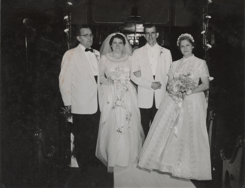 A bride and groom stand with a woman holding flowers and a man