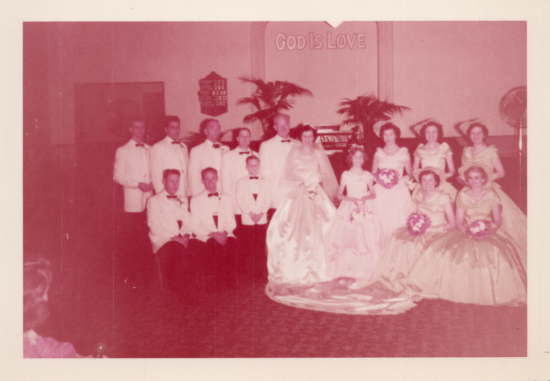 A bride and groom, surrounded by their bridesmaids and groomsmen, smile for a portrait under the text "God is Love" in the sanctuary at 19th and Green Streets