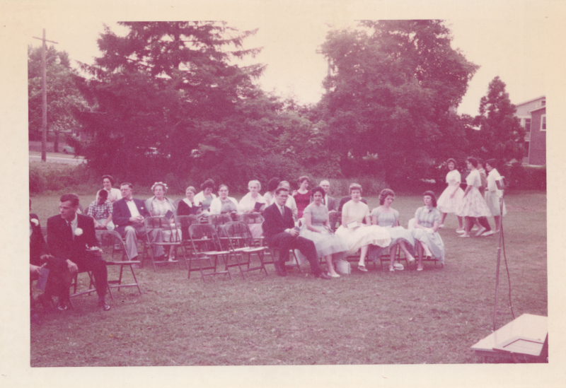 A group of formally-dressed people sit on chairs on a grass field facing an empty podium with a microphone