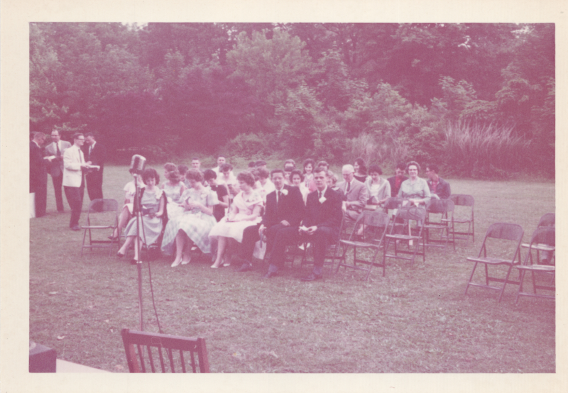A group of formally-dressed people sit on chairs on a grass field facing an empty podium with a microphone