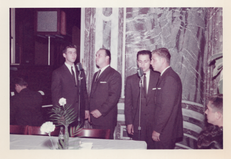 Four men sing together in front of microphones