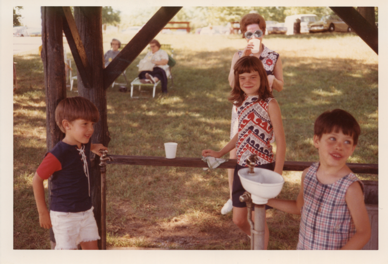 Three children smile around a water fountain outside with women in the background