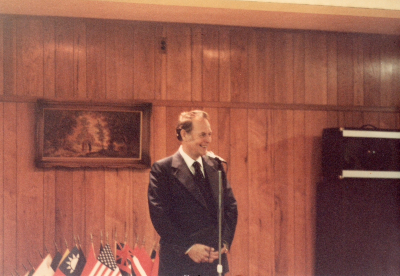 A man smiles while speaking into a microphone with world flags