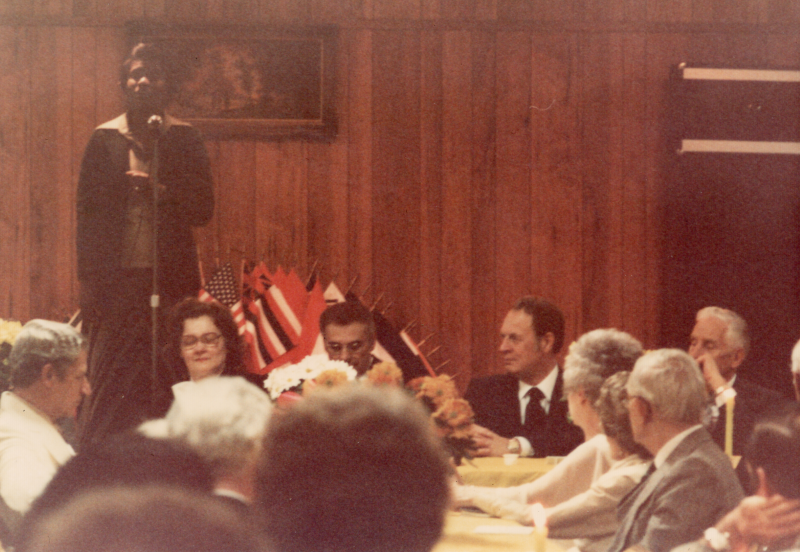 A man speaks into a microphone next to world flags facing listeners seated at tables adorned with flowers