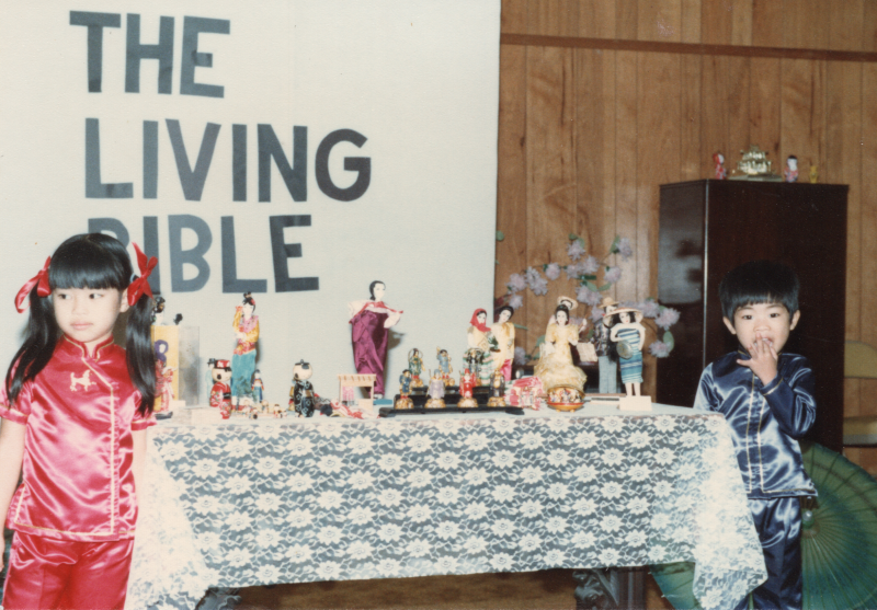 Two children stand with dolls in front of a banner that reads "The Living Bible" for Missionary Day