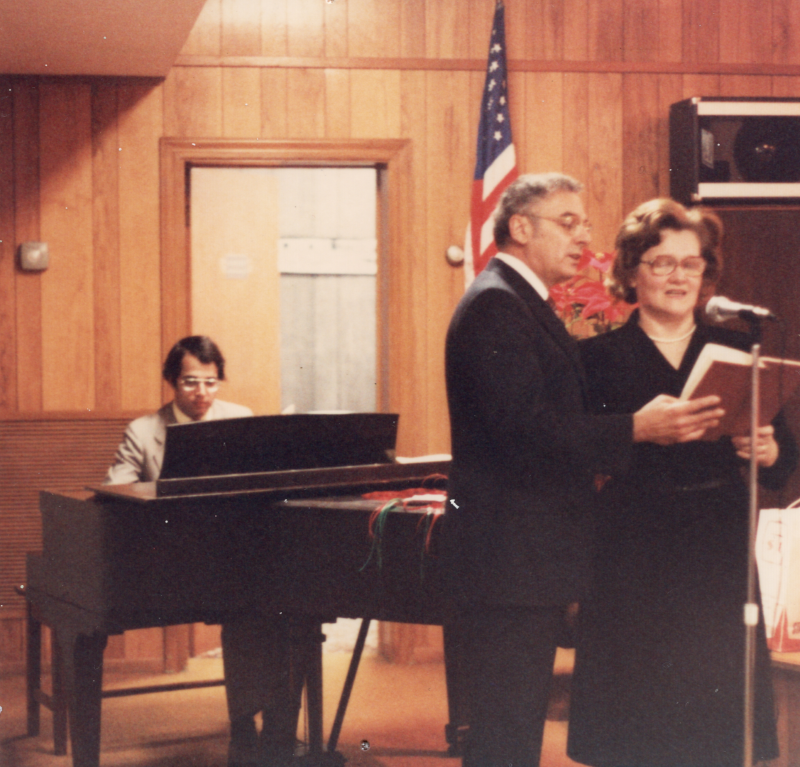 Rev. and Mrs. Bogdan sing from a hymnal in front of a microphone with a man playing piano in the background