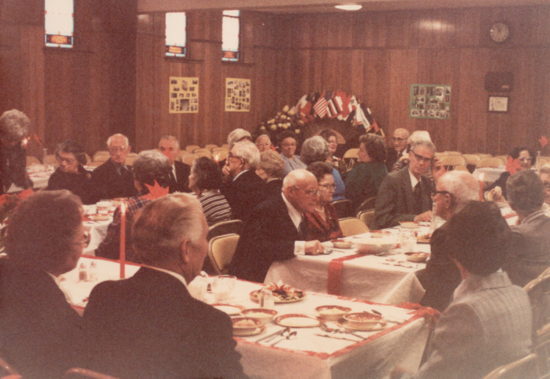 Congregants dine together around tables. World flags are in the background