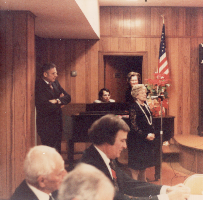 A woman sings at a microphone for congregants sitting together. Pastor and Mrs. Bogdan stand in the background with a man playing piano