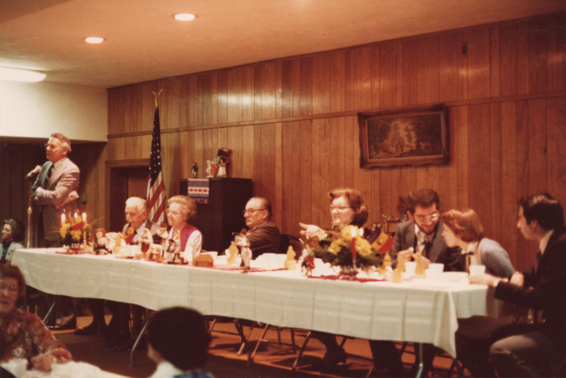 Pastor Bogdan speaks at a microphone while other congregants dine together