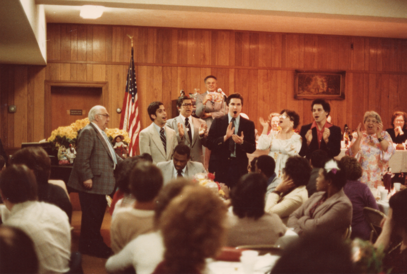 A group sings a song together in front of a room of congregants dining together