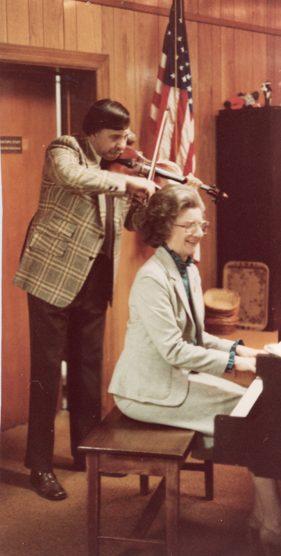 Betty Roberson playes the piano while a man stands behind her playing the violin