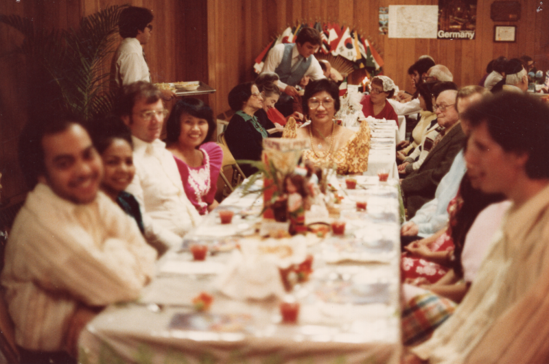 A group dines together with men serving them and international flags in the background