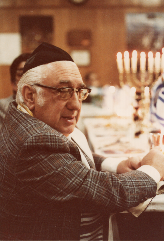A man in a yarmulke and stole is photographed with a menorah and Israeli flags at a fellowship meal
