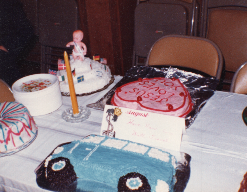 A spread of cakes including one shaped like a vehicle, one heart-shaped cake reading, "Jesus Loves You," a cake with a baby in a sash and world flags, and multicolored cakes. A sign reads "August"