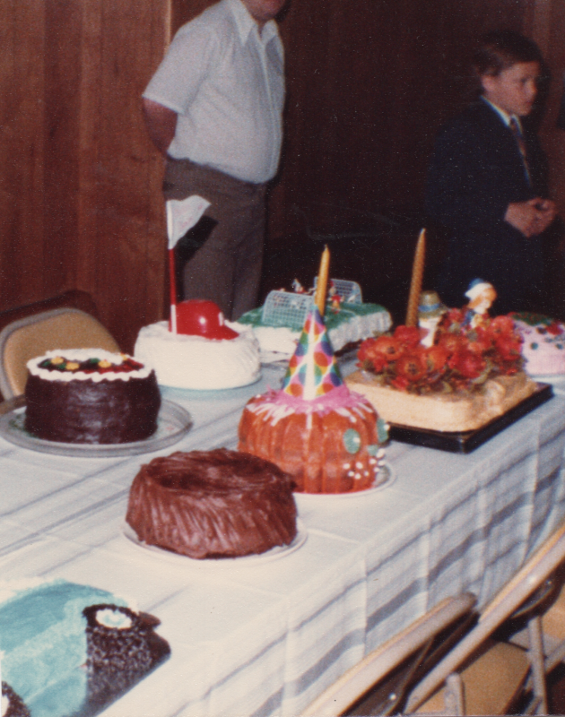 A spread of cakes including two chocolate cakes, a cake decorated as a jack-o-lantern with a party hat on, a cake with red-orange flowers, a cake with a miniature soccer game, and a cake with a flag and miniature Phillies hat.