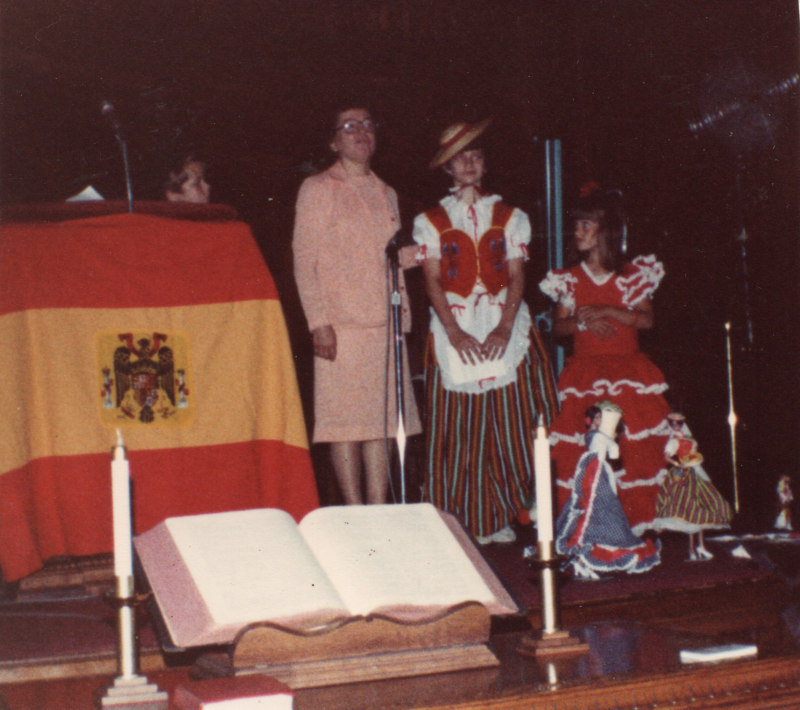 Four girls in costume perform next to a Spanish flag and two dolls in the sanctuary