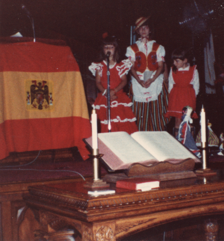 Three girls in costume perform next to a Spanish flag and two dolls in the sanctuary