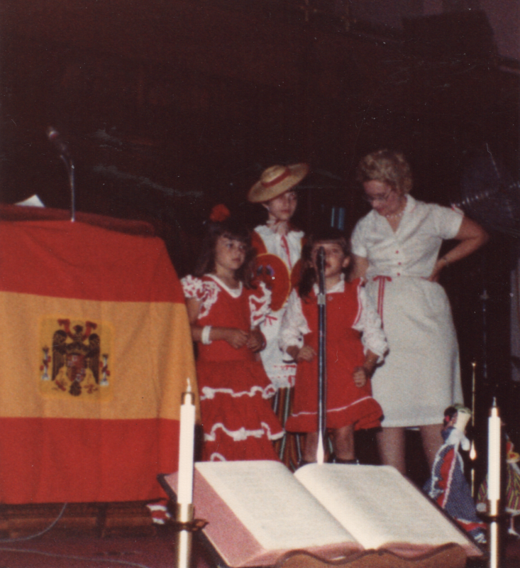 Three girls in costume perform with a woman next to a Spanish flag and two dolls in the sanctuary