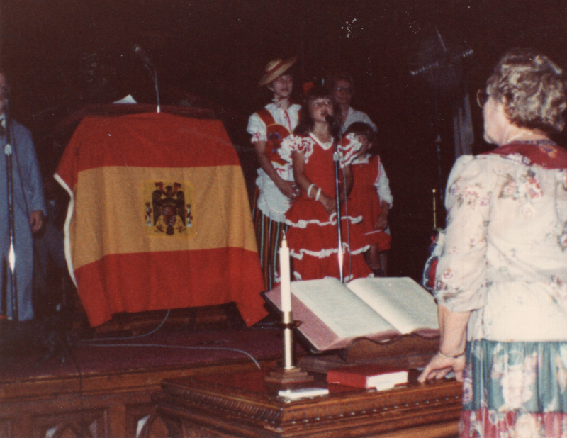 Three girls in costume perform with a woman next to a Spanish flag and two dolls in the sanctuary. A boy stands on the other side of the podium, and a woman stands in front of the stage to guide them