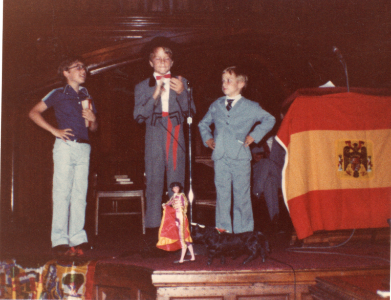 Three boys in costume perform on stage in the sanctuary next to a Spanish flage and a doll