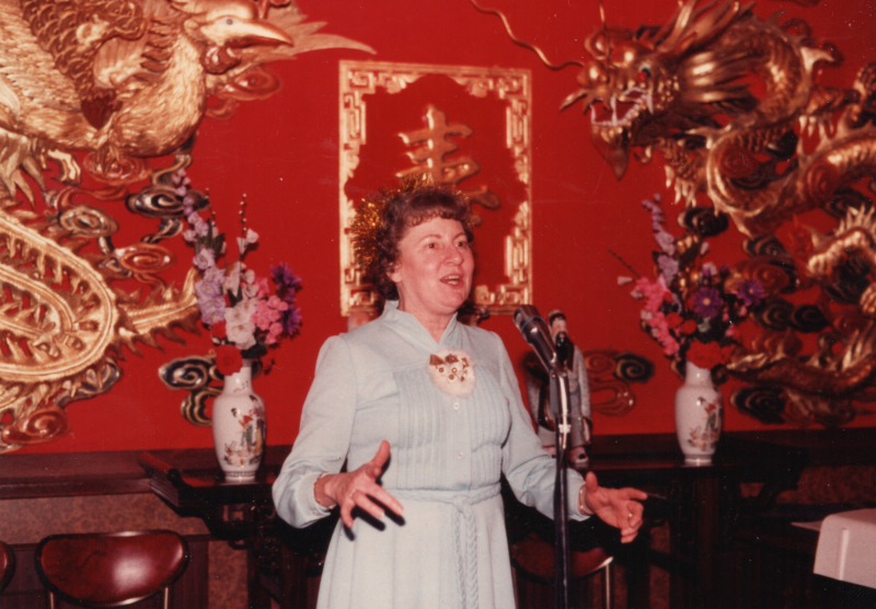 A woman in a blue dress sings into a microphone in front of a red wall adorned with gold figures